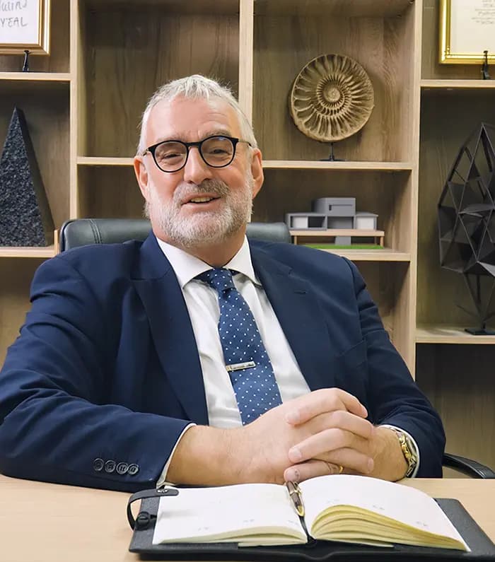 An investment advisor smiling behind a desk, with his notebook over the table and bhind him, a cabinet filled with sculptures, diplomas  and a 3d house mockuo