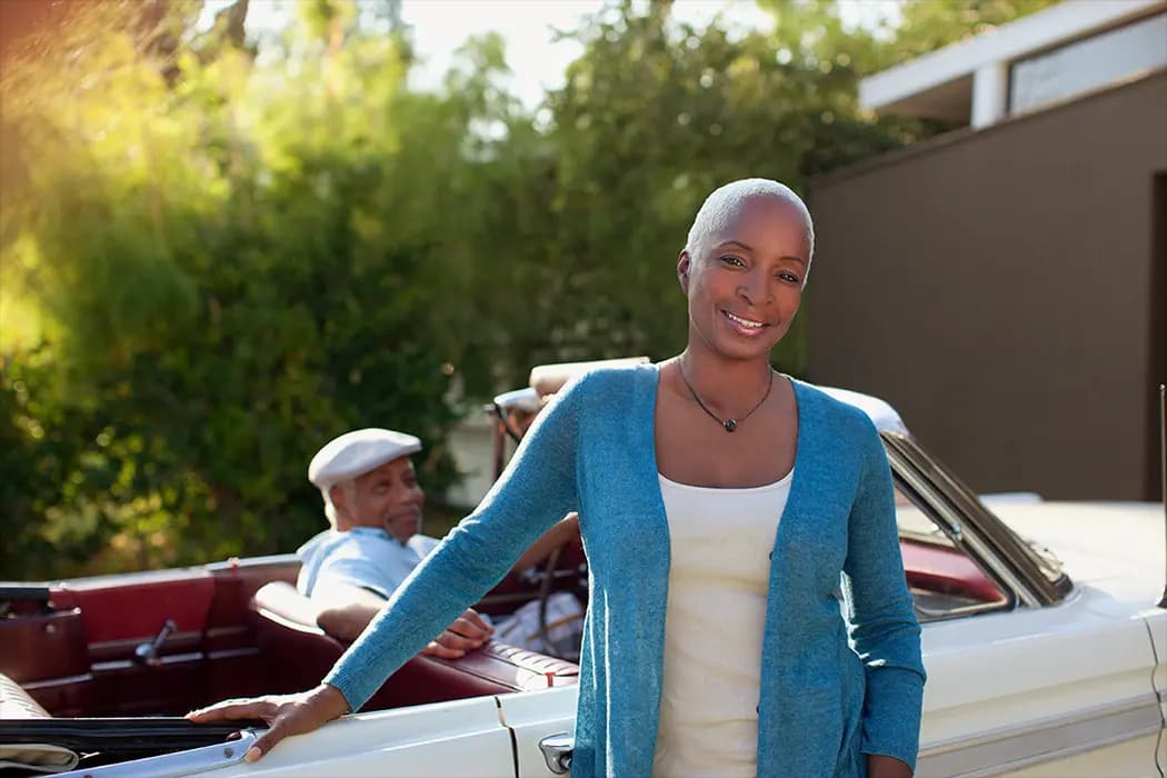 A smiling African American woman about to get into a convertible as her partner waits behind the wheel to start the journey.